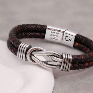 May include: A braided leather bracelet in shades of brown and red, featuring a silver-toned knot design. The bracelet has a magnetic clasp with the engraved text "Love you forever xx Anne" and two heart symbols. A stylish accessory.
