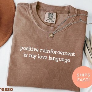 Puede incluir: Camiseta color espresso con la frase "positive reinforcement is my love language" en blanco. Un collar plateado con un colgante descansa sobre la camiseta. También se ven un sombrero marrón y hojas secas.