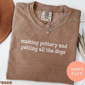 Puede incluir: Camiseta color espresso con el texto blanco "making pottery and petting all the dogs". Un collar plateado con un colgante descansa sobre la camiseta. Un sombrero marrón es visible en la esquina superior izquierda.