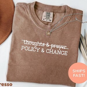 以下が含まれることがあります： エスプレッソ色のTシャツに、白で「thoughts & prayer POLICY & CHANGE」と書かれています。ペンダント付きのシルバーネックレスがシャツの上にあります。「SHIPS FAST！」と書かれたオレンジ色のステッカーが見えます。