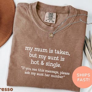 Op de afbeelding: Espresso-kleurig T-shirt met de tekst "my mum is taken, but my aunt is hot & single." Een zilveren ketting met een hanger ligt op het shirt. Het shirt heeft een label met "Comfort Colors". Een ronde sticker zegt "SHIPS FAST!"