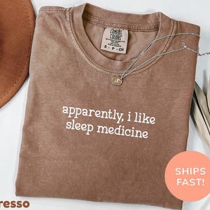 Peut inclure: Un t-shirt marron à manches courtes avec un col rond, portant l'inscription blanche "apparently, i like sleep medicine" sur le devant. Le t-shirt présente une étiquette de marque Comfort Colors au col indiquant une taille small.