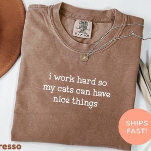 May include: Espresso-coloured t-shirt with the text "i work hard so my cats can have nice things" in white. The shirt has a crew neck and short sleeves. A silver necklace with a pendant is draped across the shirt.