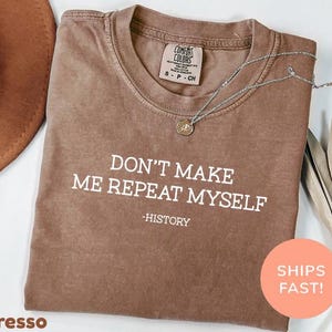 May include: Espresso-colored t-shirt with the text "DON'T MAKE ME REPEAT MYSELF - HISTORY" in white. The shirt has a crew neck and short sleeves. A silver necklace with a pendant is draped across the shirt.