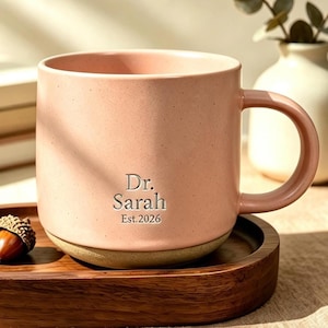 Puede incluir: Una taza de cerámica rosa claro moteada con asa, que presenta el texto grabado Dr. Sarah Est. 2026. La taza se asienta sobre una bandeja ovalada de madera marrón oscuro junto a dos pequeñas bellotas marrones con tapas texturizadas.