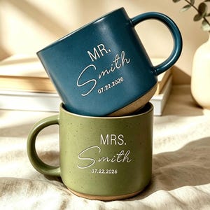 May include: A dark teal mug stacked on top of a sage green mug. Both mugs feature a speckled finish, a handle, and white printed text. The top mug reads MR. Smith 07.22.2026 and the bottom mug reads MRS. Smith 07.22.2026 in a mix of fonts.