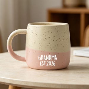 Pode incluir: Uma caneca de cerâmica de dois tons com o topo creme salpicado e a base rosa sólida. A caneca apresenta uma alça curva e o texto impresso em branco "GRANDMA EST. 2026" na frente. O design é simples, com um acabamento liso e formato arredondado.