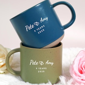 Puede incluir: Dos tazas de cerámica, una azul y otra verde, apiladas una encima de la otra. La taza azul tiene un asa oscura y el texto "Pete & Amy 9 YEARS 2025". La taza verde tiene el mismo texto. Las tazas están rodeadas de flores.
