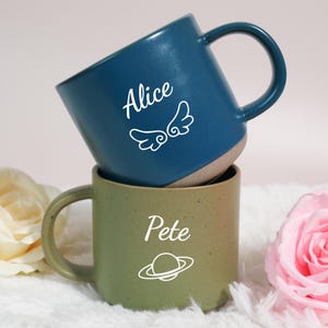 Puede incluir: Dos tazas de cerámica, una turquesa y otra verde oliva, apiladas. La taza turquesa tiene el nombre "Alice" y un diseño de alas. La taza verde oliva tiene el nombre "Pete" y un diseño de planeta. Ambas tazas tienen un asa redondeada.