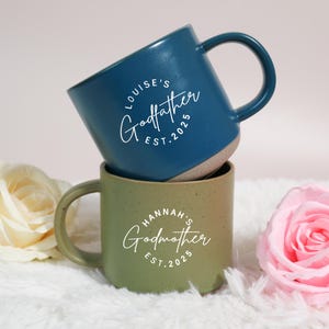 Puede incluir: Dos tazas de cerámica, una azul y otra verde, apiladas. La taza azul tiene el texto "LOUISE'S Godfather EST. 2025" en escritura blanca. La taza verde tiene "HANNAH'S Godmother EST. 2025".