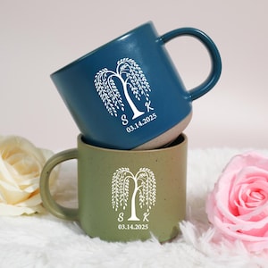 Puede incluir: Dos tazas de cerámica, una azul y otra verde, apiladas una encima de la otra. Cada taza presenta un diseño de sauce llorón blanco, las iniciales "S K" y la fecha "03.14.2025". Las tazas tienen un acabado mate y forma redondeada.