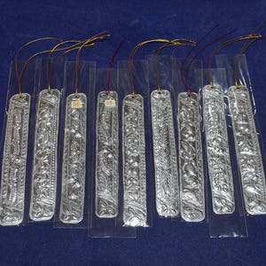 May include: A set of nine rectangular silver-colored bookmarks, each individually encased in a clear plastic sleeve. Each bookmark features an intricate embossed floral pattern and includes a thin gold-colored hanging cord at the top.