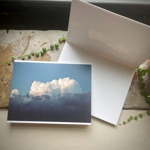 Puede incluir: Una tarjeta de felicitación blanca en blanco, abierta para revelar el interior en blanco, junto a una tarjeta con una fotografía de una formación de nubes. La imagen de la nube muestra un cielo azul con una gran nube blanca y esponjosa sobre una capa de nubes más oscuras.