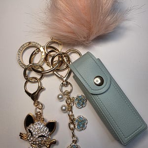 May include: A gold-coloured keychain featuring a sparkling cat charm, a pale blue case, and a fluffy pink pom-pom. The keychain also has a charm with blue flower accents and faux pearls. The cat charm is covered in clear rhinestones.