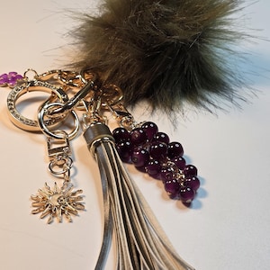 May include: A selection of keyrings with diverse charms. The keyrings include a gold-coloured ring with clear rhinestones, a gold sun charm, a grey tassel, and a cluster of purple beads. A large, fluffy, olive-green pom-pom is also attached.