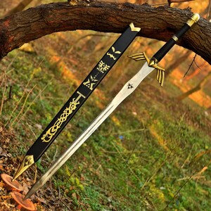 Puede incluir: Una espada plateada con empu&ntilde;adura negra y detalles dorados, apoyada sobre un lecho de hojas. La espada tiene una guarda dorada con dise&ntilde;os de alas y una vaina negra con adornos dorados. La espada mide aproximadamente 91 cm de largo.