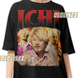 Op de afbeelding: Zwart t-shirt met een afbeelding van drie mannen met blond haar. Het woord "ICHI" is in grote rode letters bovenaan gedrukt. Het shirt heeft ook de tekst "OLDLINES" erop gedrukt.