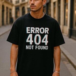 May include: Black t-shirt featuring the text "ERROR 404 NOT FOUND" in a glitch effect. The shirt is worn by a person outdoors. The text is white and teal.
