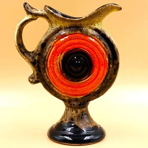 Puede incluir: Una jarra de cerámica con cuerpo circular, un centro naranja brillante y un esmalte marrón oscuro moteado. El recipiente tiene un asa curva, un pico vertedor ensanchado y una base de pedestal, mostrando un contraste marcado entre el naranja vibrante y los tonos oscuros y terrosos del acabado cerámico.
