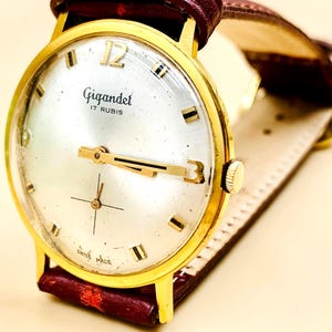 Op de afbeelding: Een vintage goudkleurig Gigandet polshorloge met een zilveren wijzerplaat, zwarte uurmarkeringen en gouden wijzers. Het horloge heeft een bordeauxrode leren band. De tekst "Gigandet 17 RUBIS SWISS MADE" is zichtbaar.