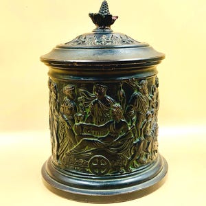 Könnte beinhalten: Ein dunkelbronze-farbener Behälter mit Deckel und einer detaillierten Reliefskulptur klassischer Figuren. Der Behälter hat einen runden Sockel und eine dekorative Spitze auf dem Deckel. Das Relief zeigt eine Szene mit Figuren in drapierter Kleidung.