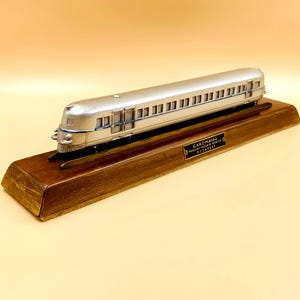 Può includere: Un modellino in metallo color argento di una carrozza ferroviaria aerodinamica montato su una base rettangolare in legno. Una piccola targhetta color ottone sulla base riporta il testo GANZ-MAVAG, MOZDONY-VAGON ES GEPGYAR, BUDAPEST in nero.