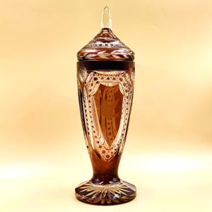 Può includere: Un alto contenitore in vetro color ambra scuro con coperchio abbinato, caratterizzato da intricati motivi in vetro tagliato, tra cui un motivo a scudo centrale con la sagoma di un uccello, un puntale appuntito e un design a stella sulla base.