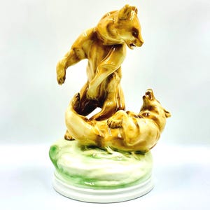 Op de afbeelding: Een keramieken beeldje van twee bruine beren in een dynamische pose. Eén beer staat boven de ander. De basis is wit met een groen en wit accent. Decoratief object.