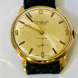 May include: Vintage gold-toned Cortébert wristwatch with a black leather band. The watch face features gold hands and markers, with the text "Cortébert 17 JEWELS INCABLOC SWISS MADE". The watch has a classic, elegant design.