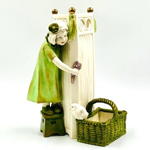 Puede incluir: Figura de cerámica de una niña con un vestido verde, sosteniendo flores, asomándose alrededor de una columna blanca. También hay una cesta verde y un taburete pequeño. La figura tiene detalles dorados y está sobre un fondo blanco.