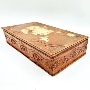 Pode incluir: Uma caixa retangular de estilo antigo com acabamento cor de cobre. A tampa apresenta um design floral com flores em relevo de cor creme. As laterais e bordas são adornadas com intrincados padrões florais esculpidos, criando uma estética vintage.