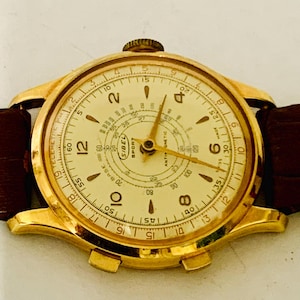 Pode incluir: Um relógio de pulso dourado com mostrador creme e pulseira de couro castanho. O mostrador apresenta algarismos pretos, ponteiros dourados e o texto "SIBEL SPORT". O relógio tem um design clássico e vintage.