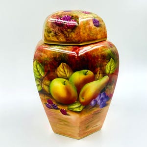 Może przedstawiać: Ozdobny ceramiczny słoik z pokrywką, z ręcznie malowanym wzorem jabłek, gruszek, winogron i jeżyn. Słoik ma kwadratową podstawę i zaokrąglony korpus, z pasującą pokrywką. Paleta kolorów obejmuje odcienie zieleni, żółci, czerwieni i fioletu.