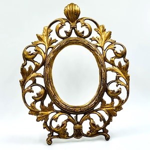 Könnte beinhalten: Ovale Bilderrahmen in Gold mit detailliertem, geschwungenem Blattdesign. Der Rahmen hat eine strukturierte ovale Mitte und ein dekoratives muschelartiges Element oben. Er steht auf einem kleinen Sockel.