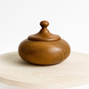 Peut inclure: Un récipient en bois artisanal avec un couvercle. Le récipient est de couleur marron chaud et de forme arrondie. Le couvercle est surmonté d'un petit bouton arrondi. Il est posé sur une surface en bois clair.