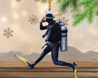 May include: A Christmas ornament featuring a scuba diver in a black and gray wetsuit, yellow fins, and a silver tank with the text "YOUR NAME." The ornament is suspended from a gold string and is set against a blurred background with snowflakes and pine branches.