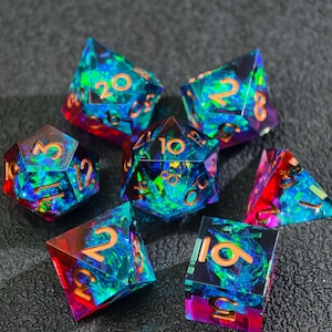 Puede incluir: Juego de siete dados poliédricos con un diseño iridiscente colorido. Los dados presentan una mezcla de tonos azules, verdes y rojos con números dorados. Los dados están diseñados para juegos de rol de mesa.
