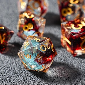May include: Close-up of several polyhedral dice. The dice are clear with gold numbering and a mix of red and blue internal designs. The dice are resting on a textured gray surface.