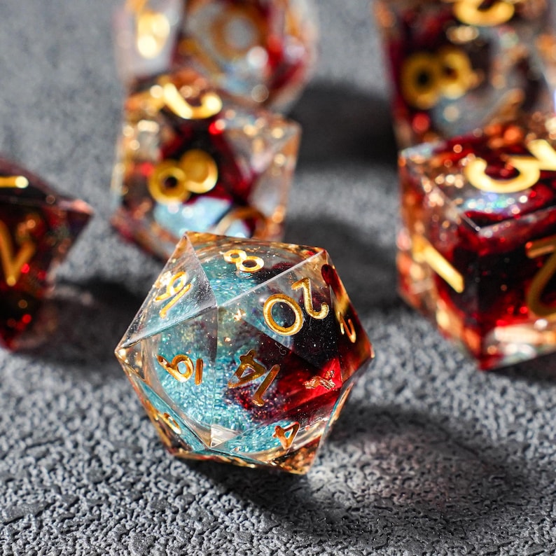 May include: Close-up of several polyhedral dice. The dice are clear with gold numbering and a mix of red and blue internal designs. The dice are resting on a textured gray surface.