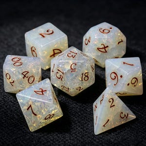 Può includere: Un set di sette dadi poliedrici realizzati in un materiale bianco latte traslucido con una texture interna iridescente e scintillante. Ogni dado presenta numeri incisi riempiti con un pigmento rosso intenso, inclusi d20, d12, d10, d8, d6, d4 e un dado percentuale.