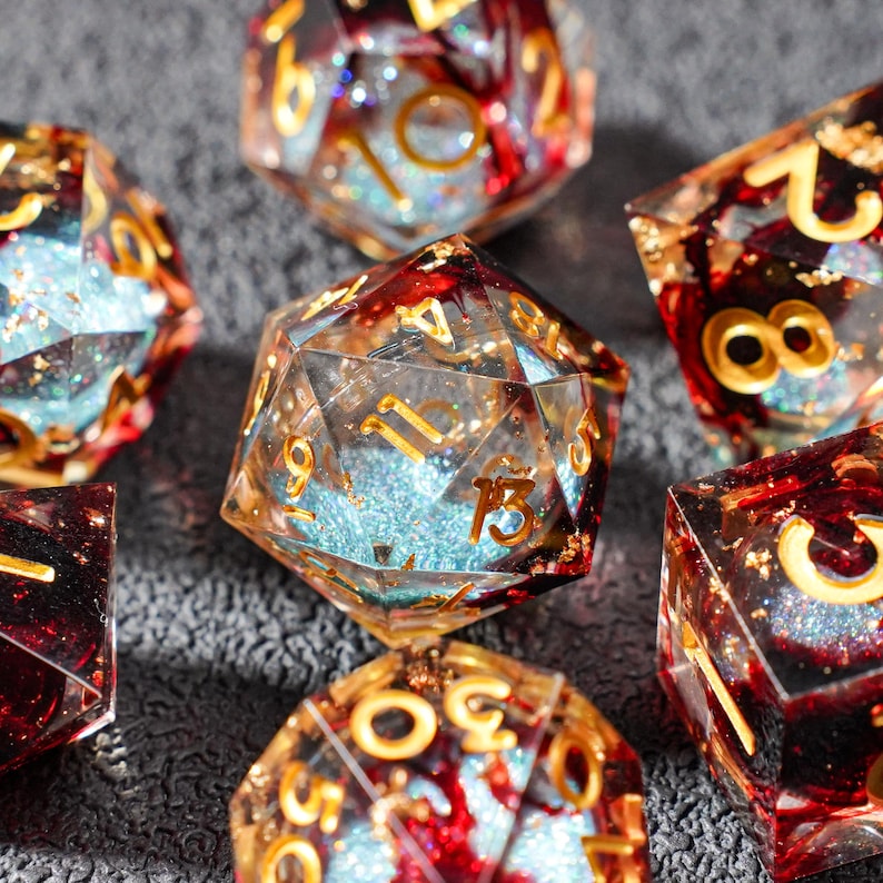 May include: A collection of polyhedral dice with a clear resin base, red and gold accents, and gold numbering. The dice have a glittery blue interior, creating a sparkling effect. The dice are designed for tabletop games.