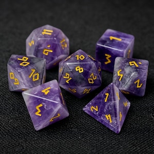Puede incluir: Un juego de siete dados poliédricos tallados en piedra amatista morada con números grabados en color dorado. La colección incluye un d20, d12, d10, d8, d6, d4 y un dado de porcentaje, todos con variaciones naturales de la piedra y bordes afilados.