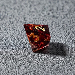 May include: A single, multi-faceted, red and gold resin die with gold numerals 2 and 3. The die is set against a textured gray surface. The die appears to be for a tabletop game.