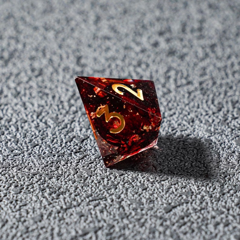 May include: A single, multi-faceted, red and gold resin die with gold numerals 2 and 3. The die is set against a textured gray surface. The die appears to be for a tabletop game.