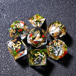 May include: A set of seven polyhedral dice featuring a skull design. Each die is clear with a skull motif, gold numbering, and red and green detailing. The dice are displayed on a dark, textured surface.