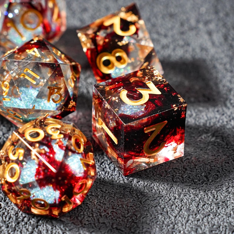 May include: A collection of polyhedral dice with a unique design. The dice are clear with red and gold accents, and gold-colored numbers. Some dice have a glittery, blue interior. The dice are designed for tabletop games.