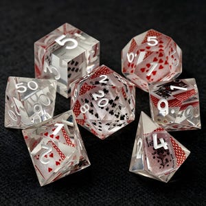 Puede incluir: Un juego de siete dados poliédricos hechos de resina transparente, cada uno con naipes en miniatura incrustados en su interior. Los dados presentan números grabados en blanco, con varios palos y valores de cartas visibles a través del material.