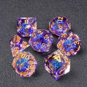 Op de afbeelding: Set van zeven polyhedrale dobbelstenen met een paars en blauw glitterontwerp. De dobbelstenen hebben goudkleurige cijfers en zijn gemaakt van een helder harsmateriaal. Deze dobbelstenen worden gebruikt voor tabletop-rollenspellen.