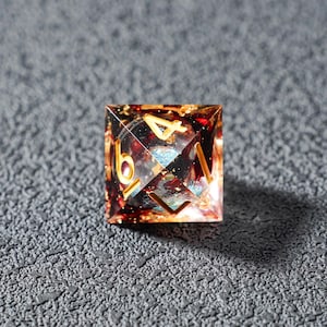 May include: A square, multi-sided die with gold numerals. The die is clear with red, gold, and blue inclusions. The numbers 4 and 6 are visible. The die is sitting on a textured, gray surface.