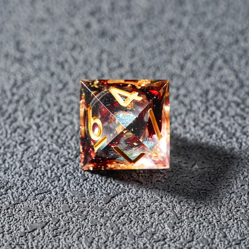 May include: A square, multi-sided die with gold numerals. The die is clear with red, gold, and blue inclusions. The numbers 4 and 6 are visible. The die is sitting on a textured, gray surface.